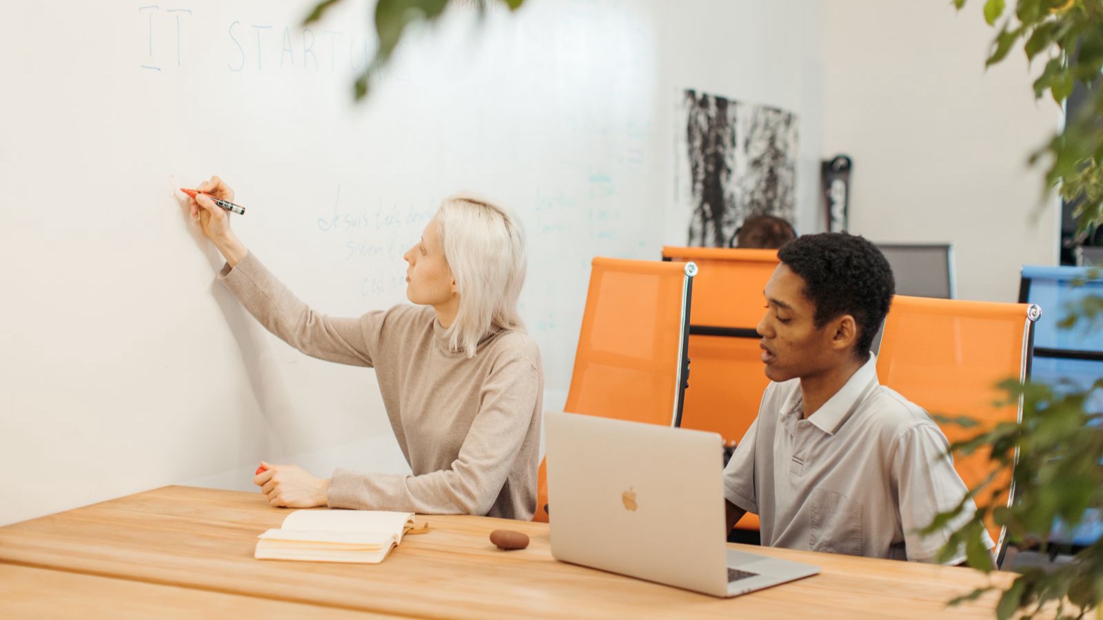 A woman writing on a whiteboard beside a man working on a laptop